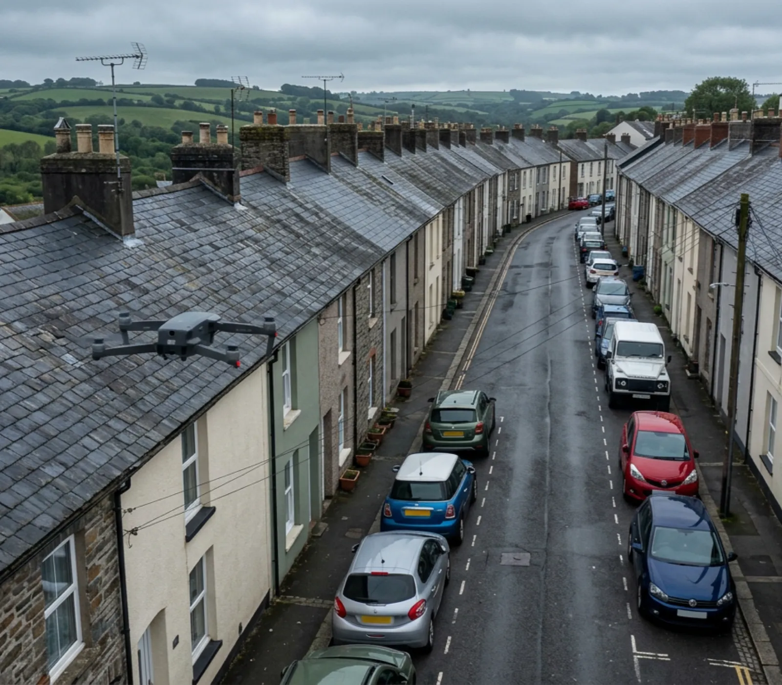 A narrow Devon terraced street with slate roofs on both sides and a small survey drone hovering above one roofline, no scaffolding or ladders in view